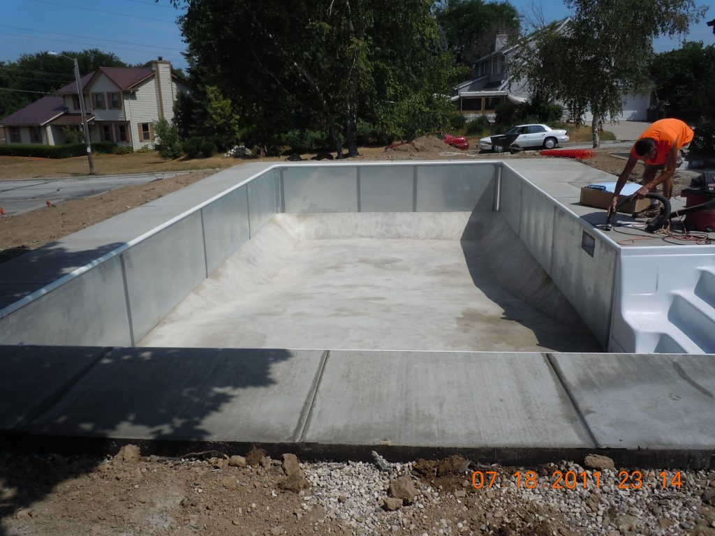 Empty rectangular in-ground pool shell during construction with concrete deck poured and a worker installing components at the pool edge.