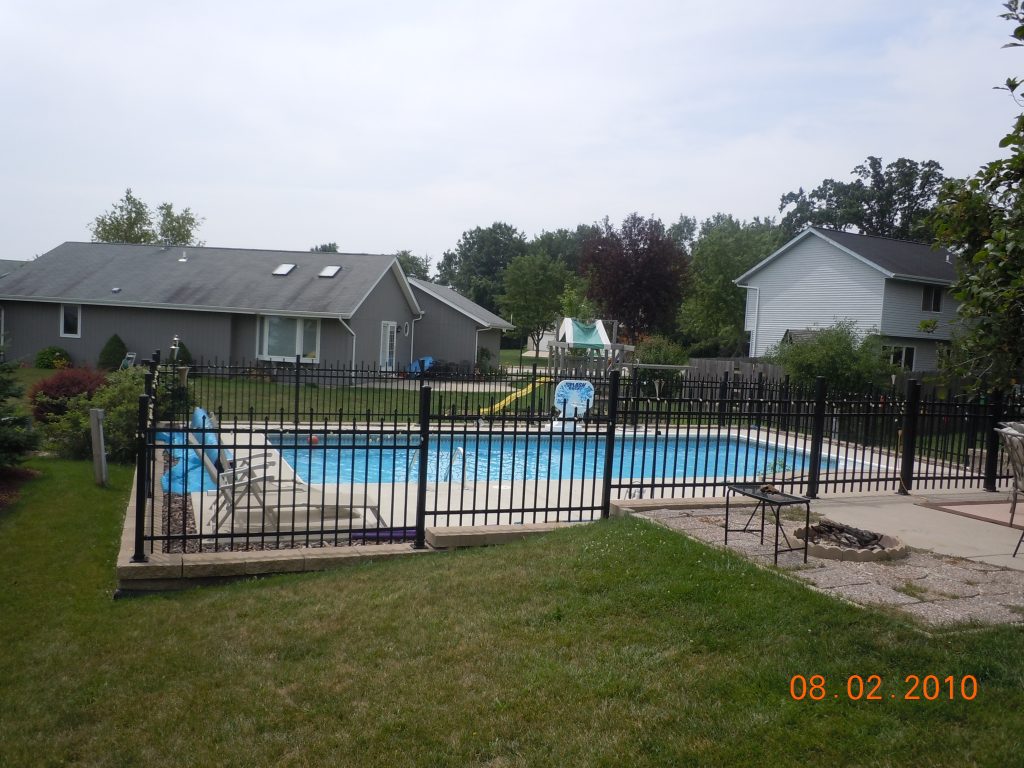 Rectangular in-ground swimming pool with blue vinyl liner, surrounded by a concrete deck and black metal fence in a residential backyard.