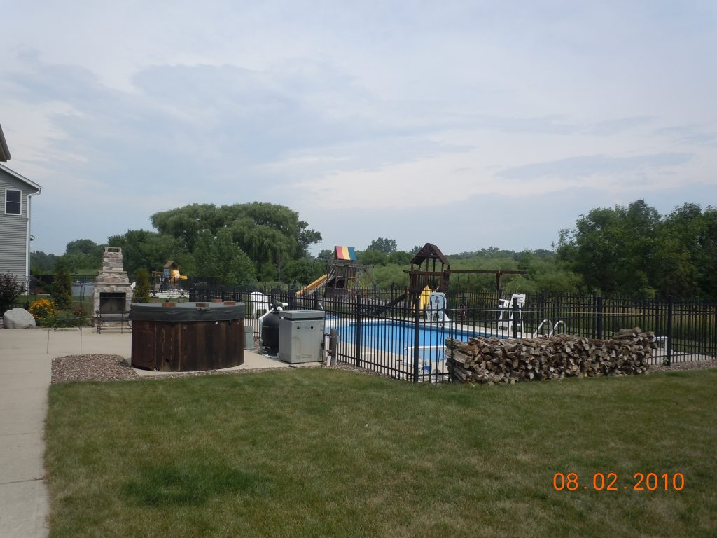 Backyard with an in-ground swimming pool behind a black metal fence, with a hot tub and pool equipment in the foreground.