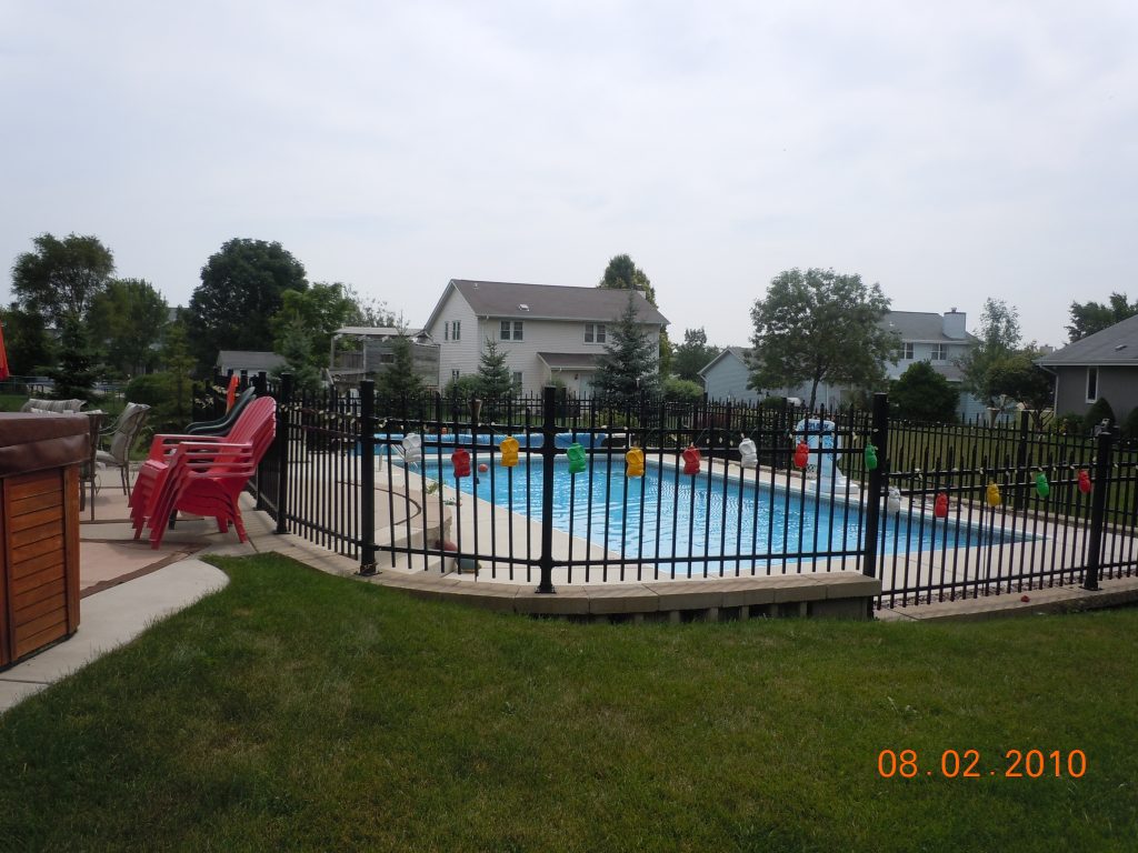 Rectangular in-ground swimming pool with concrete deck, black metal safety fence, and colorful string lights, viewed from a grassy backyard.