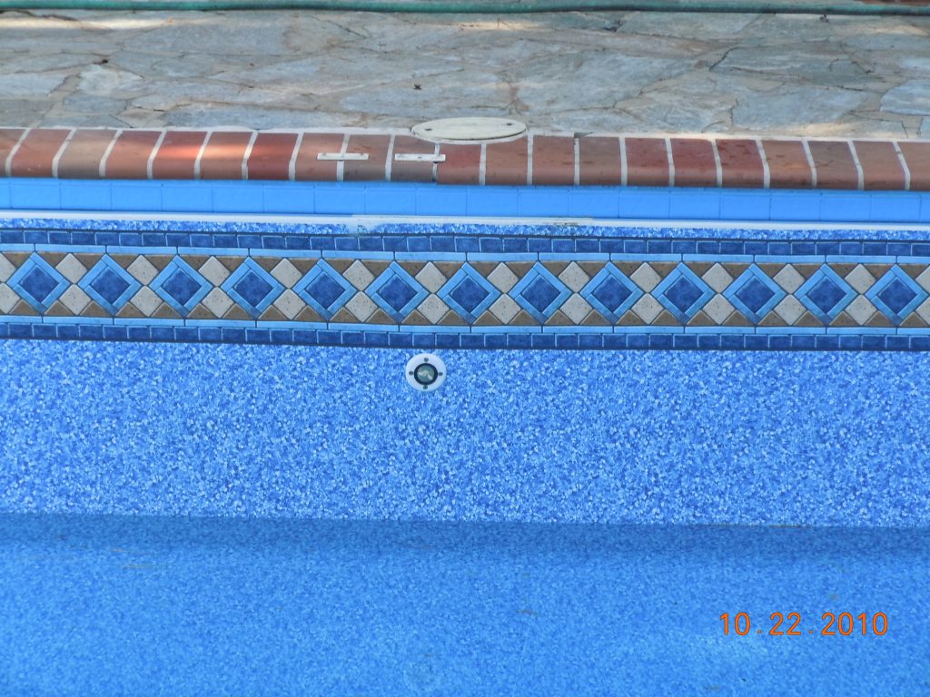 Close-up of an in-ground pool waterline showing a blue vinyl liner pattern with a decorative tile-style border beneath brick coping and a textured concrete deck.