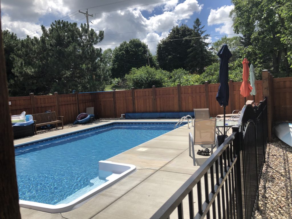 Rectangular in-ground swimming pool with blue water, concrete deck, handrail, and patio chairs along a wood privacy fence.