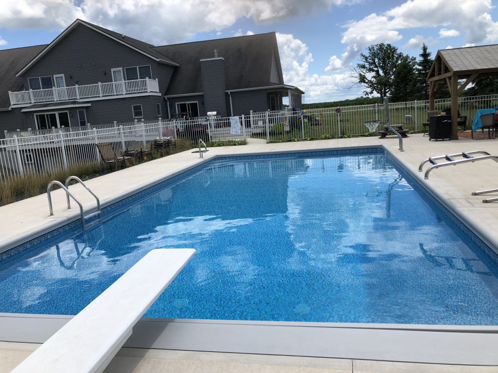 Rectangular in-ground swimming pool with blue vinyl liner, light-colored concrete deck, and handrails beside a fenced backyard.