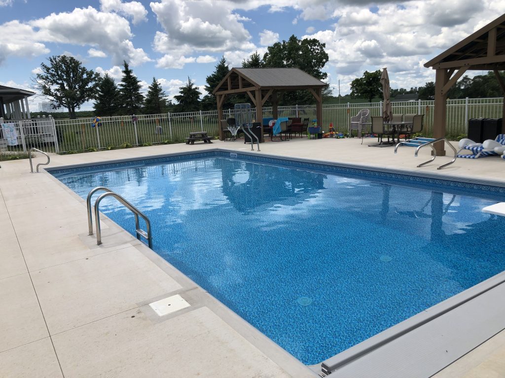Rectangular in-ground swimming pool with blue vinyl liner, concrete deck, and handrails, with a fenced backyard and patio seating in the background.