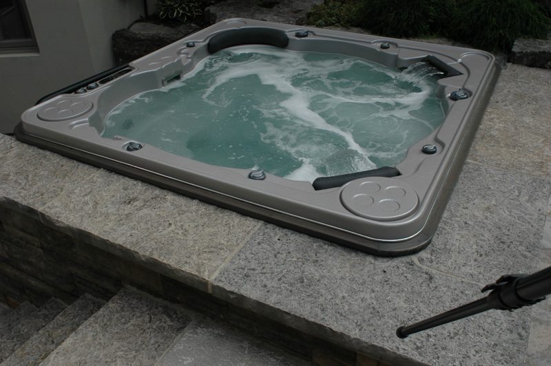 Built-in square hot tub with bubbling water set into a stone patio next to a house.