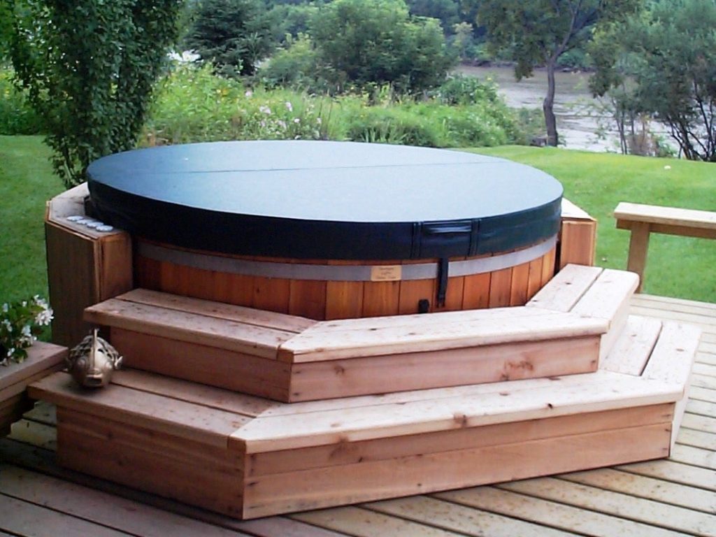 Round above-ground spa with a dark insulated cover on a tiered wooden deck in a grassy backyard.