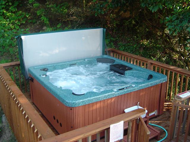 Hot tub with cover open and jets running on a wooden backyard deck surrounded by trees.