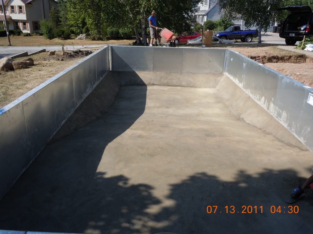 Rectangular in-ground pool shell under construction with metal wall panels and a concrete base in a residential backyard.