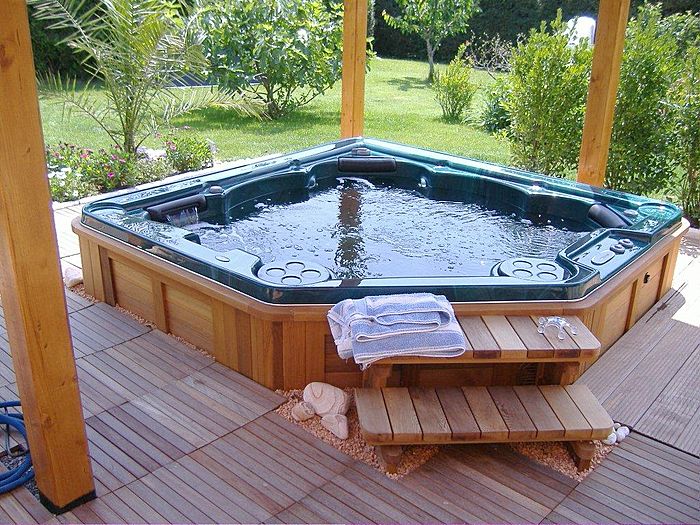 Octagonal above-ground hot tub on a wooden deck with steps, towels, and a backyard lawn in the background.