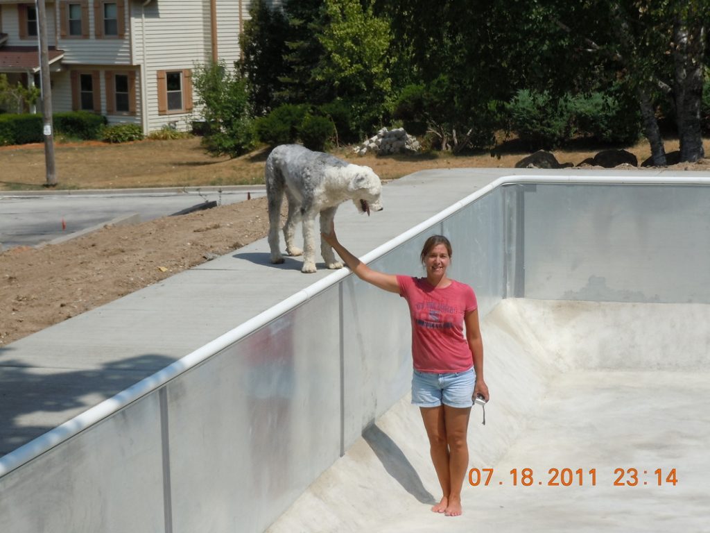 Person standing inside an empty in-ground pool during installation, reaching up to pet a dog on the pool edge.