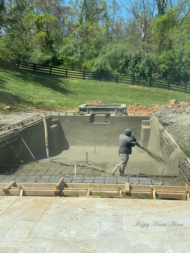 Crew member spraying gunite onto the walls of an in-ground pool shell during construction, with rebar and plumbing visible.