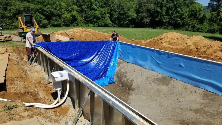 Workers position a blue vinyl liner inside a rectangular in-ground pool shell during construction, with soil piles and an excavator in the background.