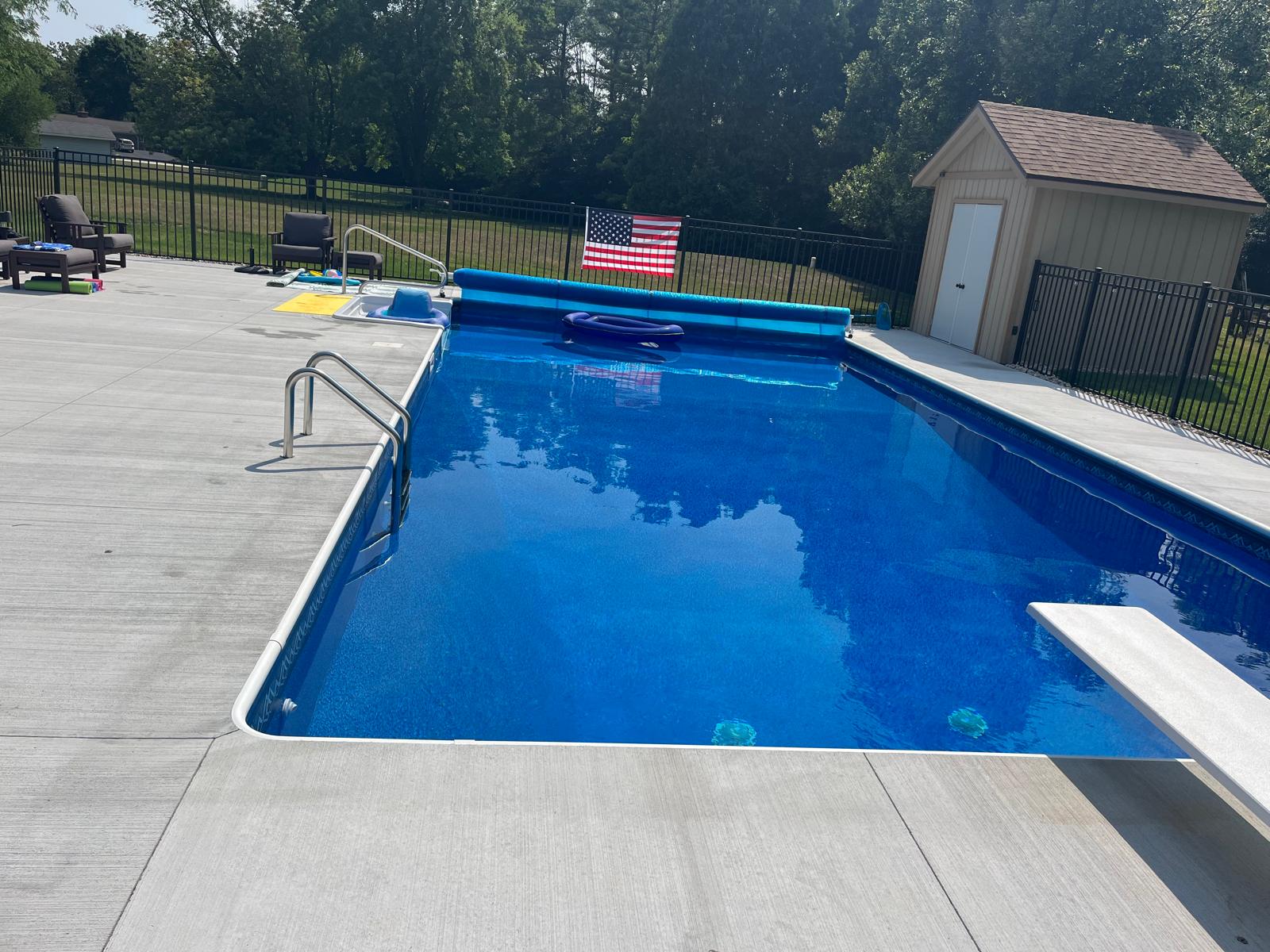 Rectangular in-ground pool with bright blue liner, concrete deck, handrails, and a diving board in a fenced backyard.