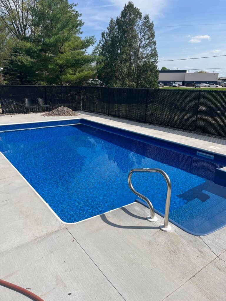 Rectangular in-ground vinyl liner pool with blue patterned liner, concrete deck, and stainless handrail on a sunny day.