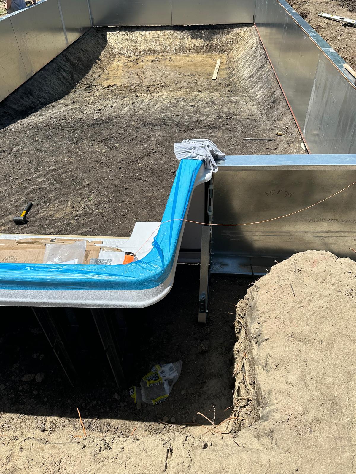 Rectangular in-ground pool under construction with steel wall panels and a white step section wrapped in blue protective plastic.