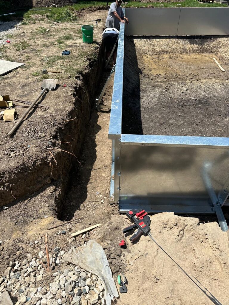Workers align a metal wall panel for a rectangular in-ground pool during construction, with the excavated trench and tools visible.