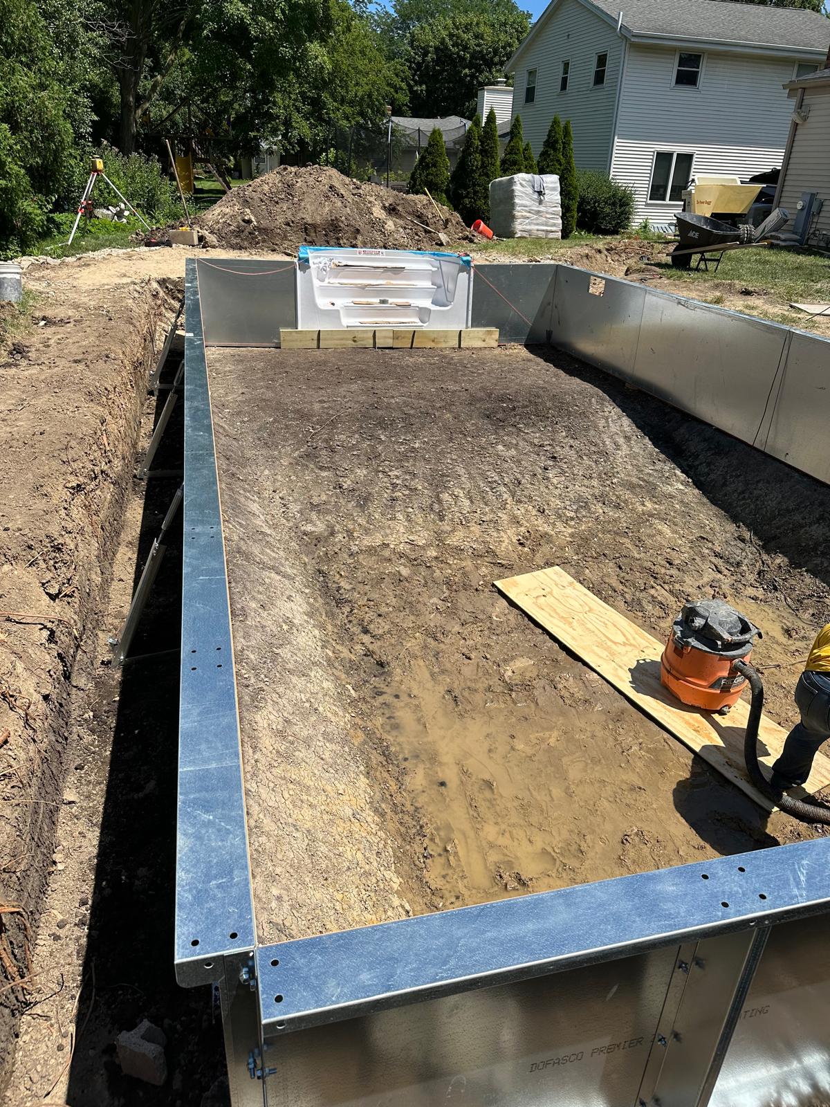 In-ground rectangular pool under construction with metal wall panels installed and white entry steps set in place.