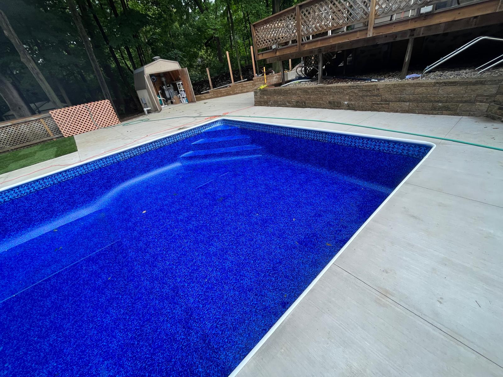 Empty rectangular in-ground pool with a bright blue vinyl liner and patterned waterline, viewed from the deck with steps visible at one end.