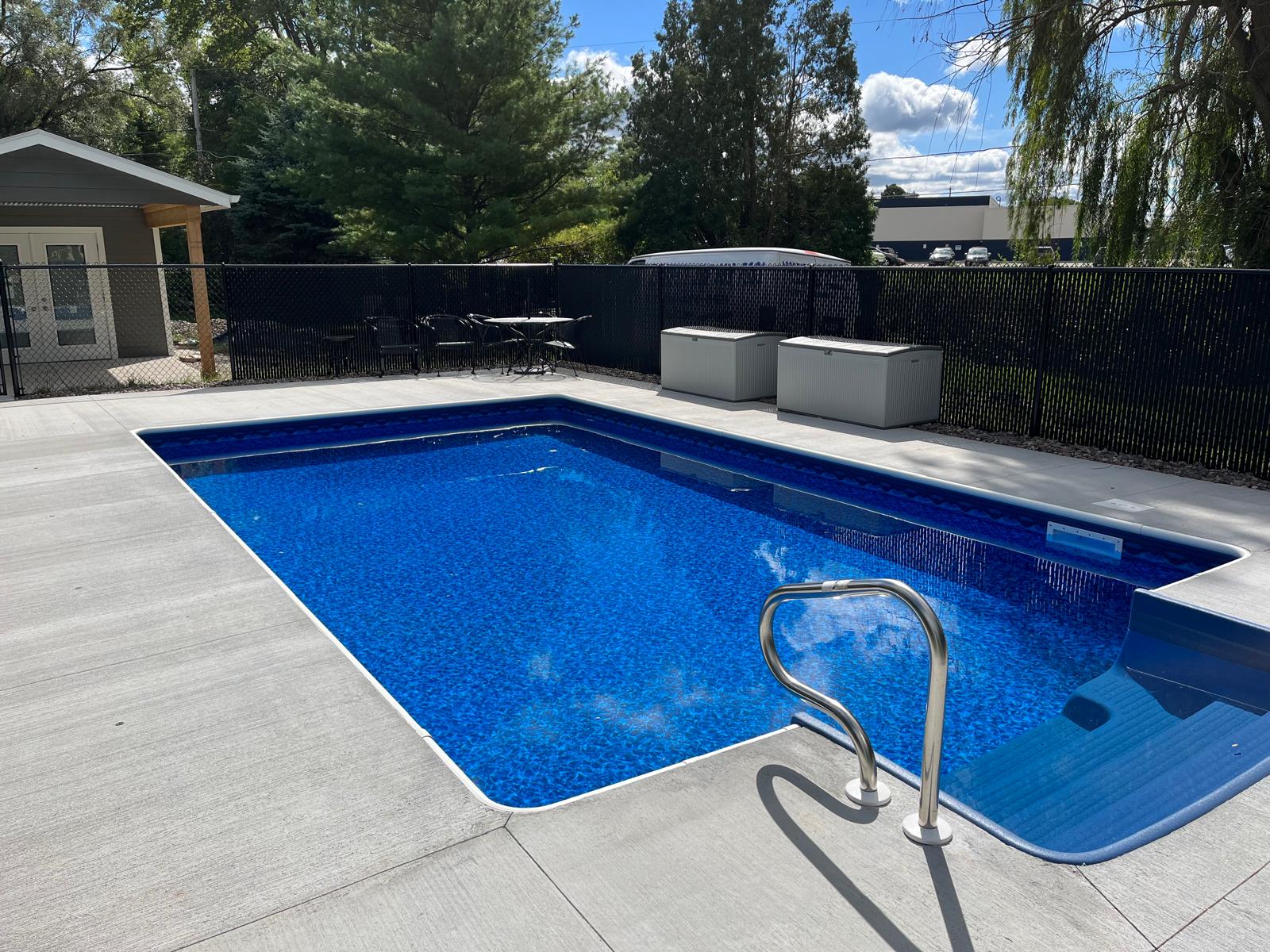 Rectangular in-ground vinyl liner pool with bright blue water, gray concrete deck, handrail, and wide entry steps in a fenced backyard.