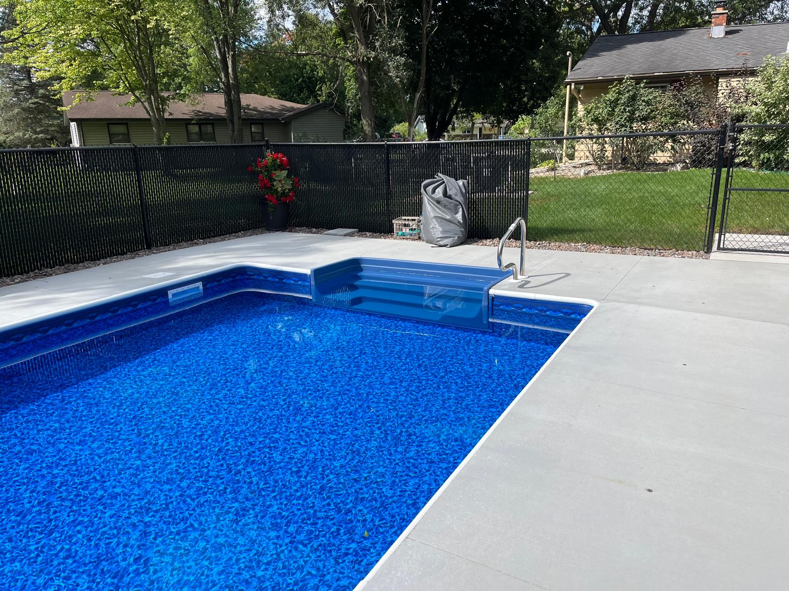 In-ground swimming pool with a bright blue vinyl liner, wide entry steps, and a light gray concrete deck in a fenced backyard.