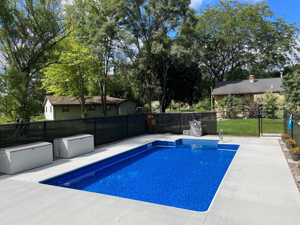 Rectangular in-ground pool with bright blue vinyl liner, wide entry steps, and a light-colored concrete deck in a fenced backyard.