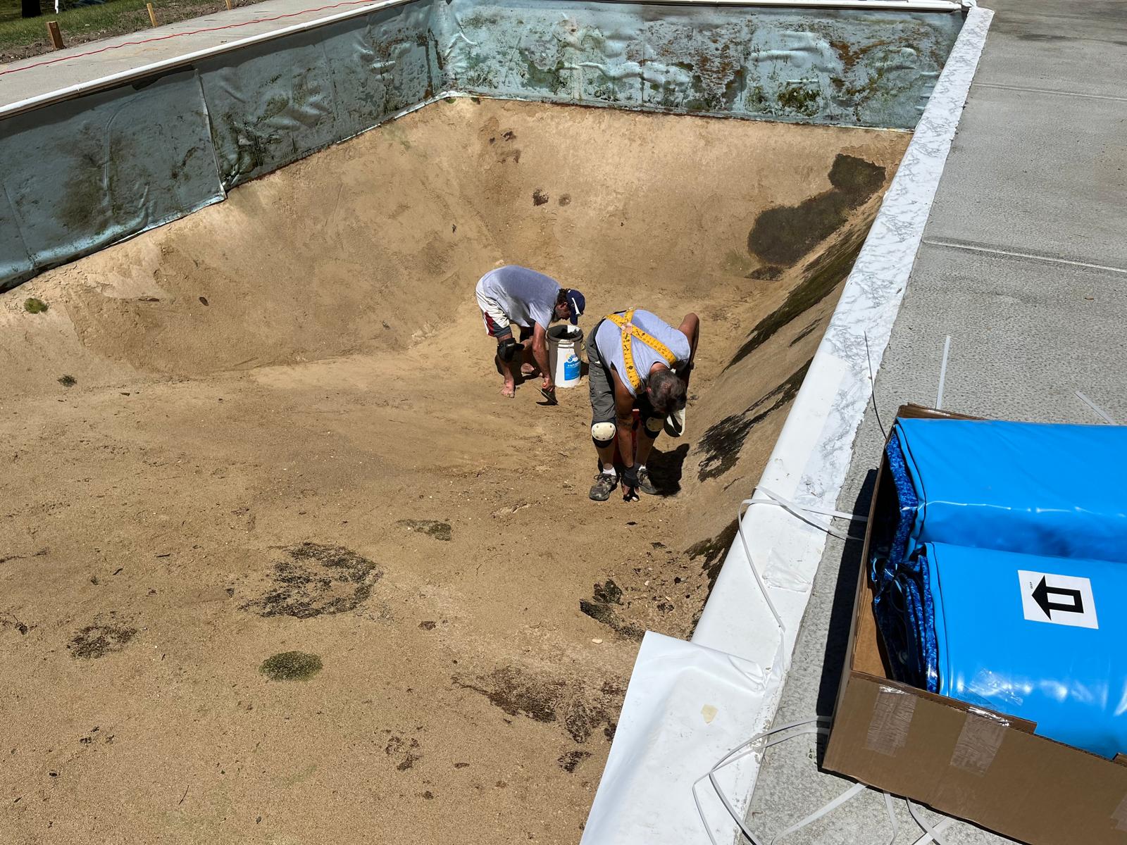 Two workers smooth the sand base inside an empty in-ground pool during liner installation.