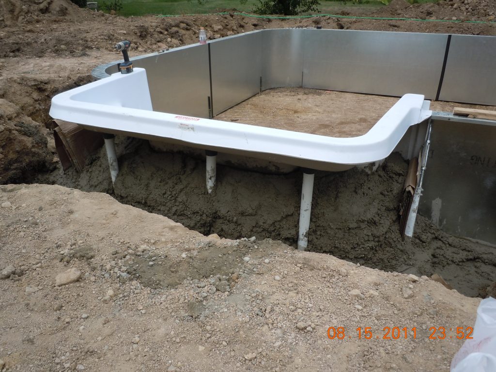 White fiberglass pool step entry installed during an in-ground pool build, with metal wall panels and excavated soil visible.